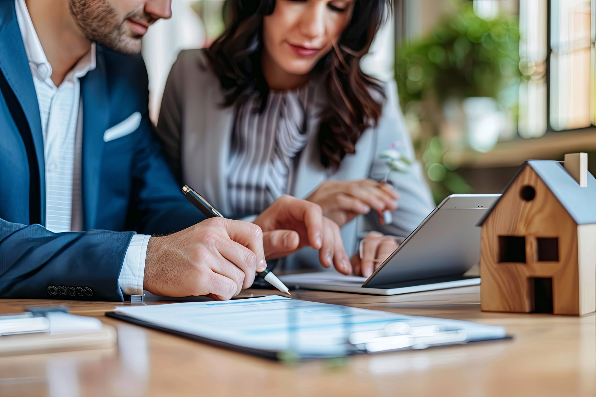 Mann im Anzug und Frau betrachten Immobilienunterlagen mit Laptop und Holzhaus-Modell auf dem Tisch