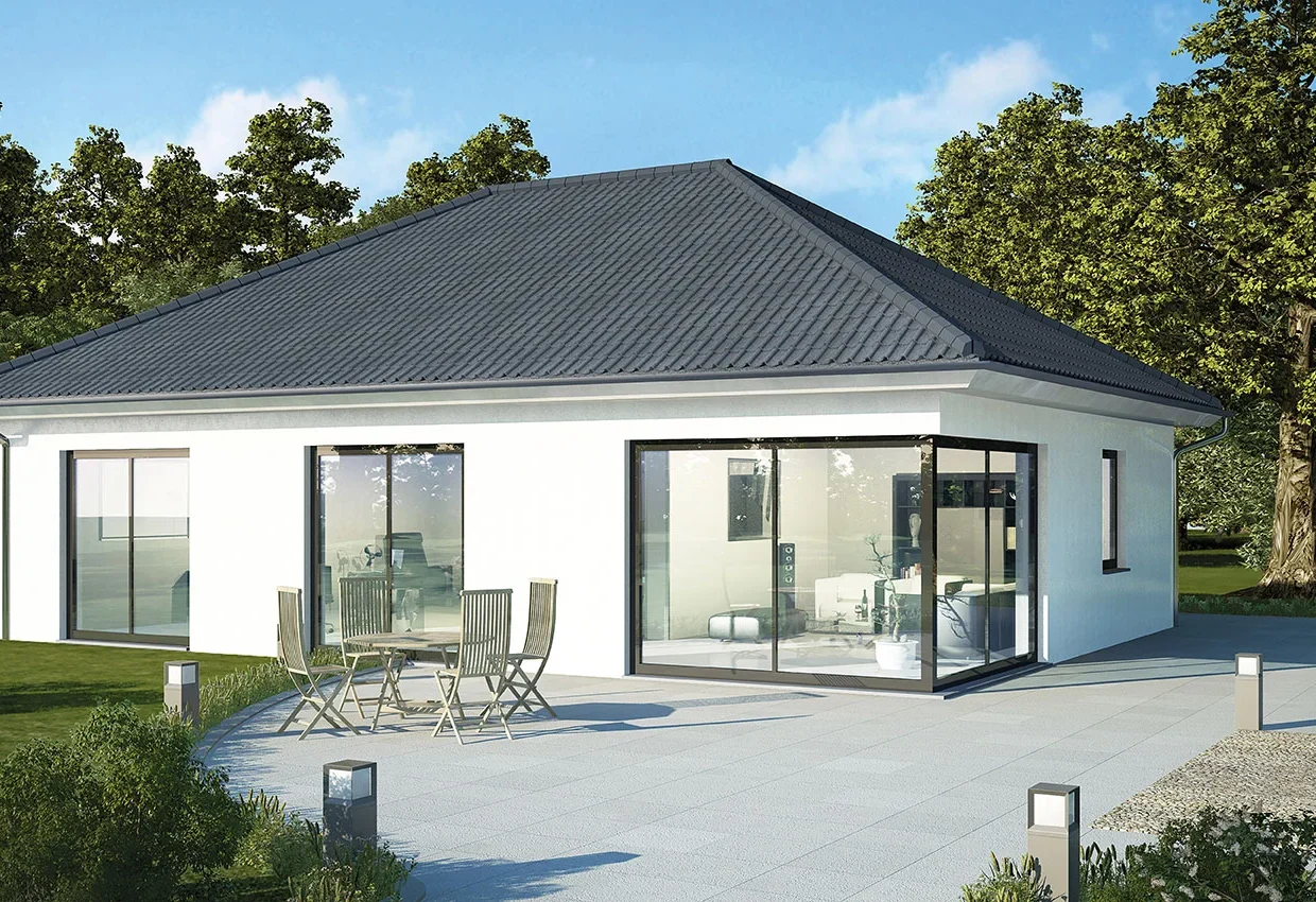 Moderner weißer Bungalow mit grauem Walmdach, großen Fenstern und Terrasse mit Gartenmöbeln in parkähnlicher Landschaft